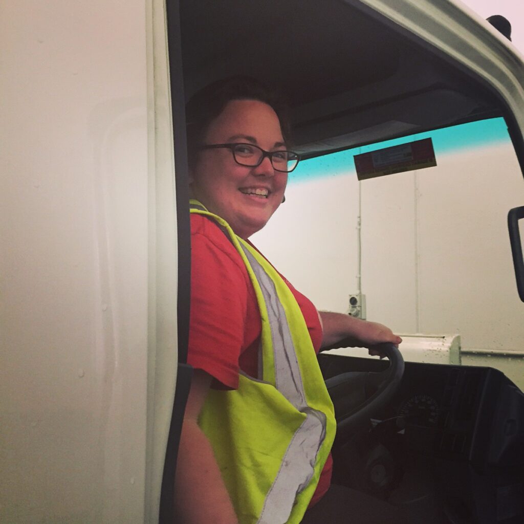 Janelle Baird smiling from driver's seat of delivery truck wearing high-visibility safety vest and glasses, in-the-trenches credibility and anti-hustle energy