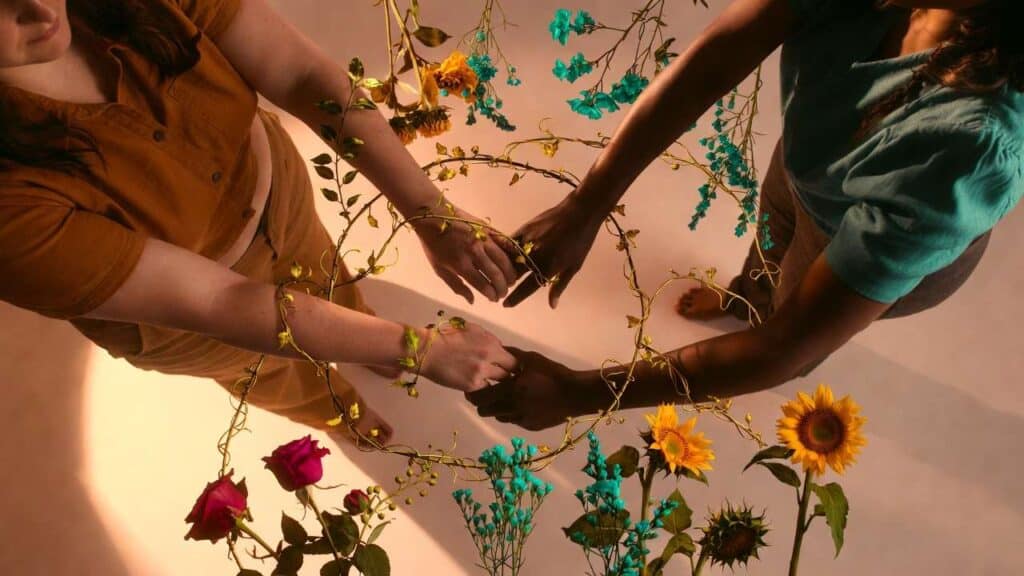 Three women of diverse body types and ethnicities in circle with hands joined in center, connected by golden and teal botanical vines, each surrounded by different flowers representing the unique support they offer